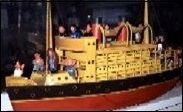 A photo of a stamped metal steam ship approximately 15 inches long. The superstructure with its two smoke stacks is yellow, the hull is painted black above the yellow waterline and red below it. On the deck are many Homies toys from the Playground series. Stretched between cargo masts at the front and rear of the deck superstructure are half a dozen Buddhist prayer flags. There are two lifeboats hanging on davits on either side of the smoke stacks.