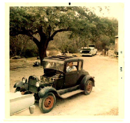 Photo of the curator sitting in a 1929 Model A coupe. The car is as it was found and is being towed from behind a creekside house to his home.