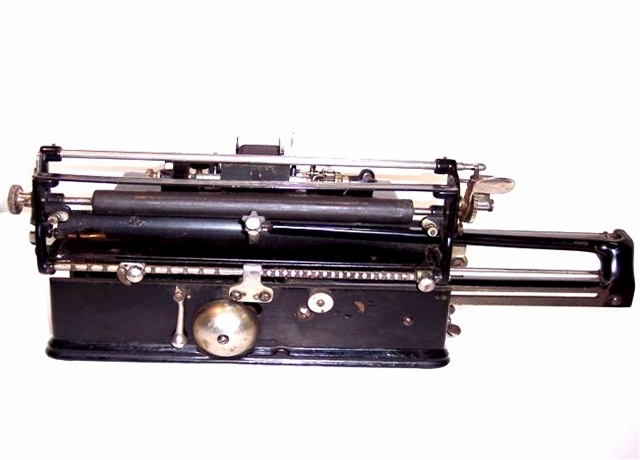 Here we see the Chicago from the rear. The carriage is still extended. The star wheel that locks the carriage down is shown here in the center of the carriage frame.To the right of it is a knurled adjustment knob.Below it is the ringer and its hammer. There are two margin locks, one at each end. For a better and more detailed account, reference patent number 