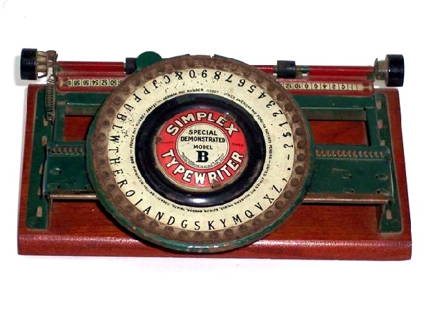 Simplex Special Demonstrated Model B Typewriter, all caps, green metal body with red metal paper feed assembly attached. The base is a dark stained wood. Two rubber paper feed rollers and two carriage twirlers. The large green dial has a black inner ring around the logo plate. The logo plate has a red ring with the words SIMPLEX TYPEWRITER in sans serif caps. Inside the red ring, centered against a white circle, are the words SPECIAL DEMONSTRATED MODEL B. All the text is sans serif except the B, which is black and in caps. The much larger B is a large, bold, black serif B.